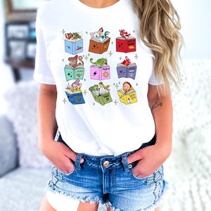 May include: White t-shirt featuring a colorful graphic of open books with animated characters. The books are in various colors, and the characters include a monkey, a lizard, and a dwarf. The design is centered on the shirt.