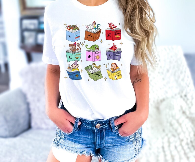 May include: White t-shirt featuring a colorful graphic of open books with animated characters. The books are in various colors, and the characters include a monkey, a lizard, and a dwarf. The design is centered on the shirt.