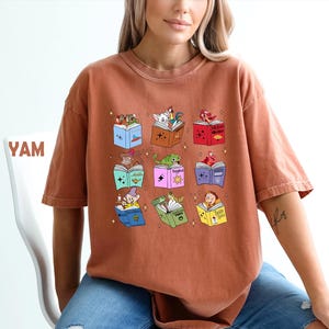 May include: Rust-colored t-shirt featuring a colorful graphic of open books with various cartoon characters. The books are in different colors and have stars around them. The word "YAM" is visible on the left.