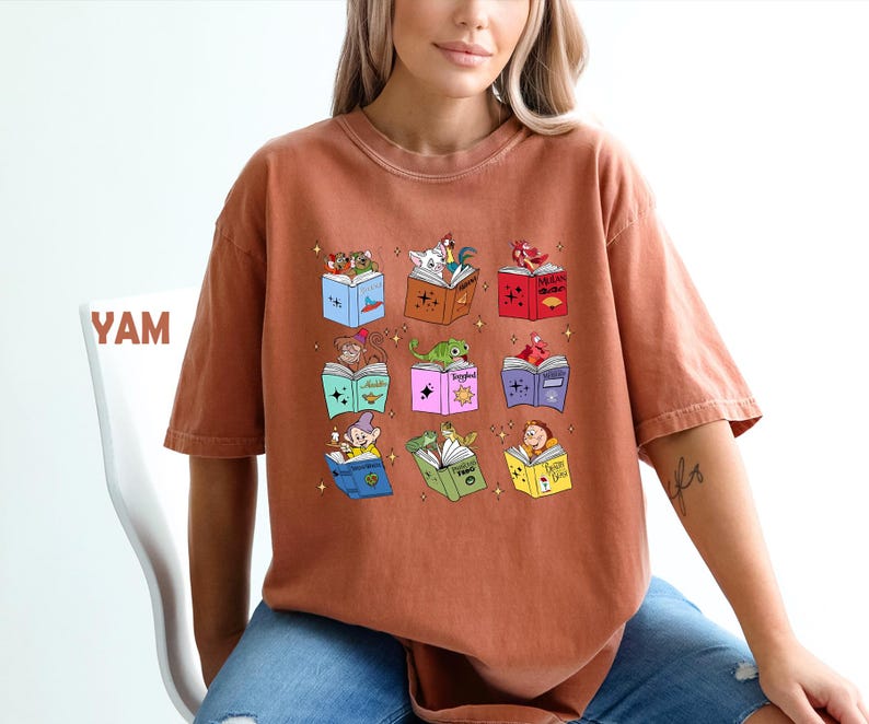 May include: Rust-colored t-shirt featuring a colorful graphic of open books with various cartoon characters. The books are in different colors and have stars around them. The word "YAM" is visible on the left.