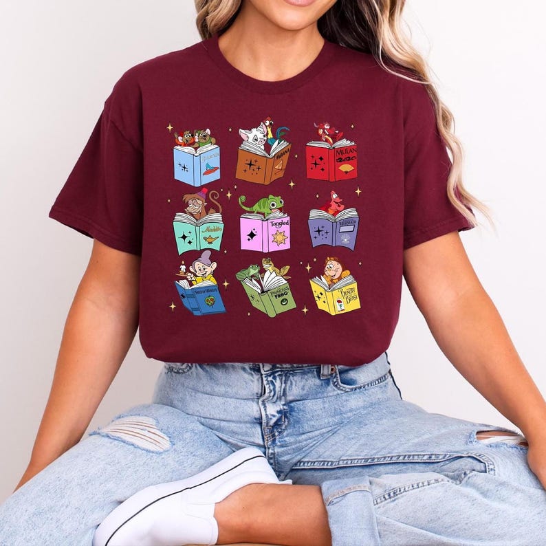 May include: Burgundy t-shirt featuring a grid of colorful storybook illustrations. Each book has a different character, including a monkey, a dwarf, and a frog. The books are open, and the characters are reading. The shirt is a casual, comfortable style.