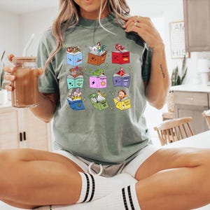 May include: Sage green t-shirt featuring a colorful graphic of open books with animated characters. The books are in various colors, and the characters include a rooster, a princess, a frog, and a dwarf. The shirt is paired with white shorts and striped socks.