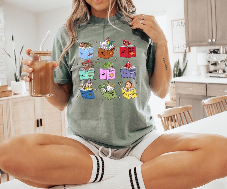 May include: Sage green t-shirt featuring a colorful graphic of open books with animated characters. The books are in various colors, and the characters include a rooster, a princess, a frog, and a dwarf. The shirt is paired with white shorts and striped socks.