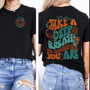 May include: Black t-shirt with a circular design on the front that says "Take a deep breath and remember who you are." The back of the shirt has the same phrase in a larger, colorful design. The design includes orange, teal, and yellow colors.