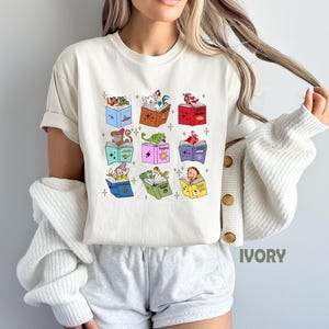 May include: Ivory-colored t-shirt featuring colorful illustrations of storybook characters reading books. The design includes a variety of characters and books in different colors. The shirt is a casual, short-sleeved style.