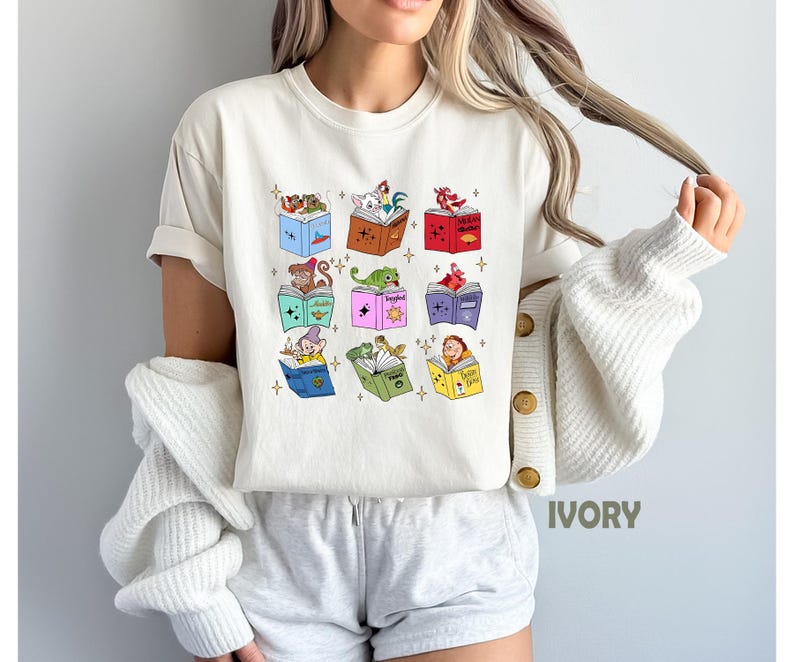 May include: Ivory-colored t-shirt featuring colorful illustrations of storybook characters reading books. The design includes a variety of characters and books in different colors. The shirt is a casual, short-sleeved style.