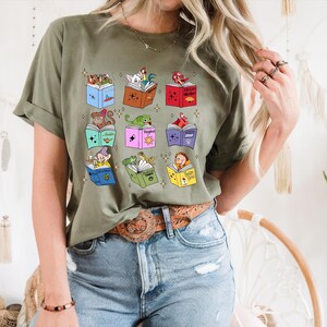 May include: Olive green t-shirt featuring a colorful graphic of open books, each with a different cartoon character. The books are in various colors, and the characters include a monkey, a dragon, and a dwarf.
