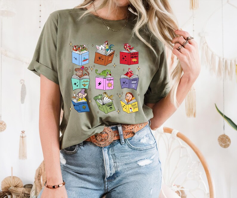 May include: Olive green t-shirt featuring a colorful graphic of open books, each with a different cartoon character. The books are in various colors, and the characters include a monkey, a dragon, and a dwarf.