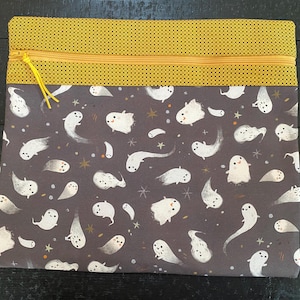 May include: A rectangular zippered pouch with a Halloween theme. The bottom section features a dark gray fabric with white ghost illustrations and gold stars. The top section is yellow with small black dots. The zipper is yellow.