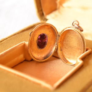 Puede incluir: Un relicario ovalado de oro abierto que presenta una piedra preciosa ovalada de color p&uacute;rpura intenso engastada en el lado interior izquierdo. El relicario tiene un dise&ntilde;o con bisagras y una peque&ntilde;a anilla met&aacute;lica en la parte superior.