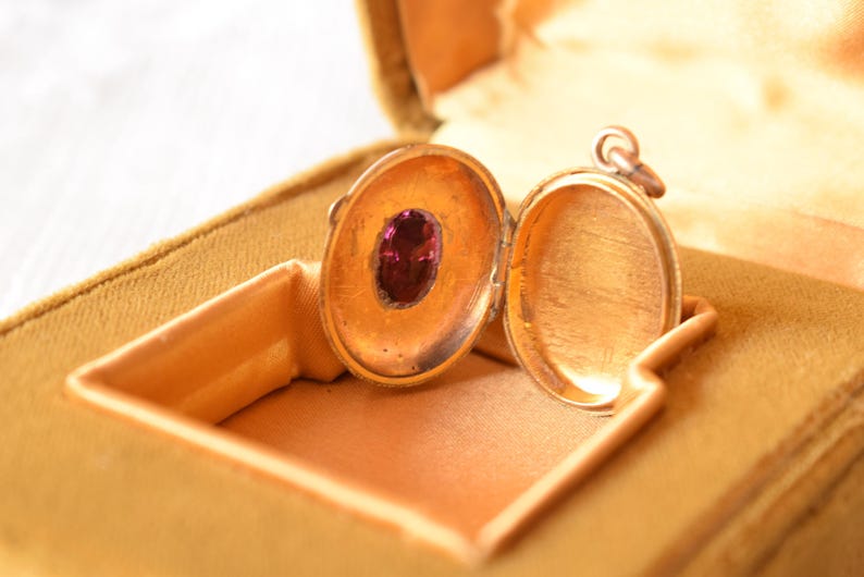 Puede incluir: Un relicario ovalado de oro abierto que presenta una piedra preciosa ovalada de color p&uacute;rpura intenso engastada en el lado interior izquierdo. El relicario tiene un dise&ntilde;o con bisagras y una peque&ntilde;a anilla met&aacute;lica en la parte superior.