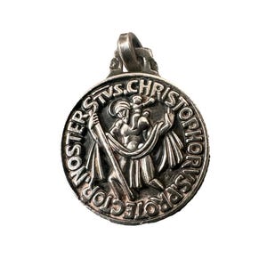 May include: A silver-colored pendant featuring a detailed depiction of Saint Christopher carrying the Christ Child. The pendant has the inscription "NOSTER STVS. CHRISTOPHORVS PROTECTOR" around the edge. The pendant has a loop at the top for a chain.