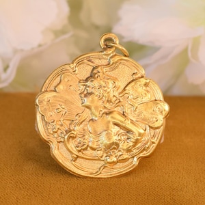 May include: A circular gold-toned pendant featuring a raised relief design of a figure with flowing hair and wings, surrounded by floral motifs. The pendant has a scalloped edge and a small metal loop at the top for attachment to a chain or cord.