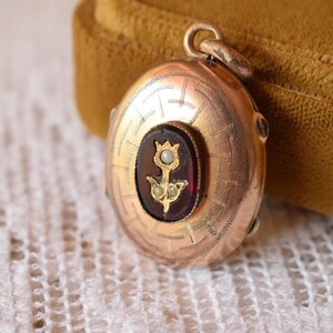 Puede incluir: Un relicario redondo de metal en tono dorado con un patr&oacute;n geom&eacute;trico grabado en la superficie. El centro muestra una incrustaci&oacute;n de piedra ovalada de color rojo oscuro con un motivo floral dorado acentuado por tres peque&ntilde;as perlas blancas.