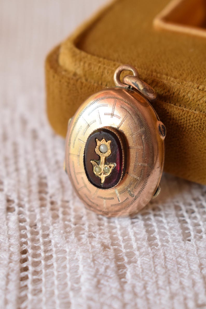 Puede incluir: Un relicario redondo de metal en tono dorado con un patr&oacute;n geom&eacute;trico grabado en la superficie. El centro muestra una incrustaci&oacute;n de piedra ovalada de color rojo oscuro con un motivo floral dorado acentuado por tres peque&ntilde;as perlas blancas.
