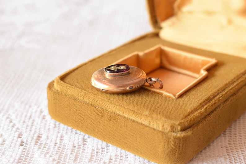 Puede incluir: Un relicario redondo de metal en tono dorado con una peque&ntilde;a incrustaci&oacute;n ovalada oscura en el centro con un delicado dise&ntilde;o floral. El relicario incluye una peque&ntilde;a anilla de metal para colgar y se presenta dentro de un joyero forrado de terciopelo color mostaza.