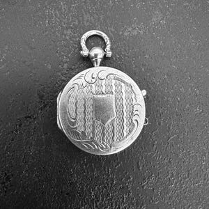 May include: A round silver-coloured metal locket featuring an intricate engraved pattern of wavy vertical lines and decorative scrollwork. The centre displays a smooth, shield-shaped cartouche. A small, ornate circular bail is attached to the top.
