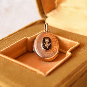 Puede incluir: Un relicario ovalado de oro con un patr&oacute;n geom&eacute;trico grabado en la superficie. El centro muestra una incrustaci&oacute;n ovalada oscura con un motivo floral dorado acentuado por dos peque&ntilde;as perlas blancas. El relicario incluye una argolla superior.