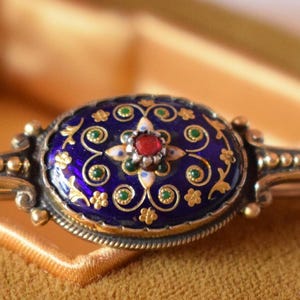 Pode incluir: Um broche de prata com uma peça central oval de esmalte azul profundo, filigrana floral dourada, pequenos detalhes verdes e uma pedra vermelha central. A moldura de metal tem uma textura perolada e dois braços horizontais polidos.