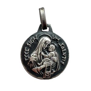 May include: A silver-toned pendant featuring a religious image of a woman holding a child. The pendant has the words "ECCE SIGNUM SALVTIS" around the edge. The pendant has a loop at the top for a chain.