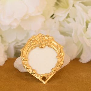 Könnte beinhalten: Ein herzförmiger, goldfarbener Metallrahmen mit einer verzierten, geprägten Bordüre aus Blumen- und Schnörkelmustern. Die Mitte des Rahmens besteht aus einer leeren, weißen, glatten Fläche, und das Stück hat eine spitze Basis.