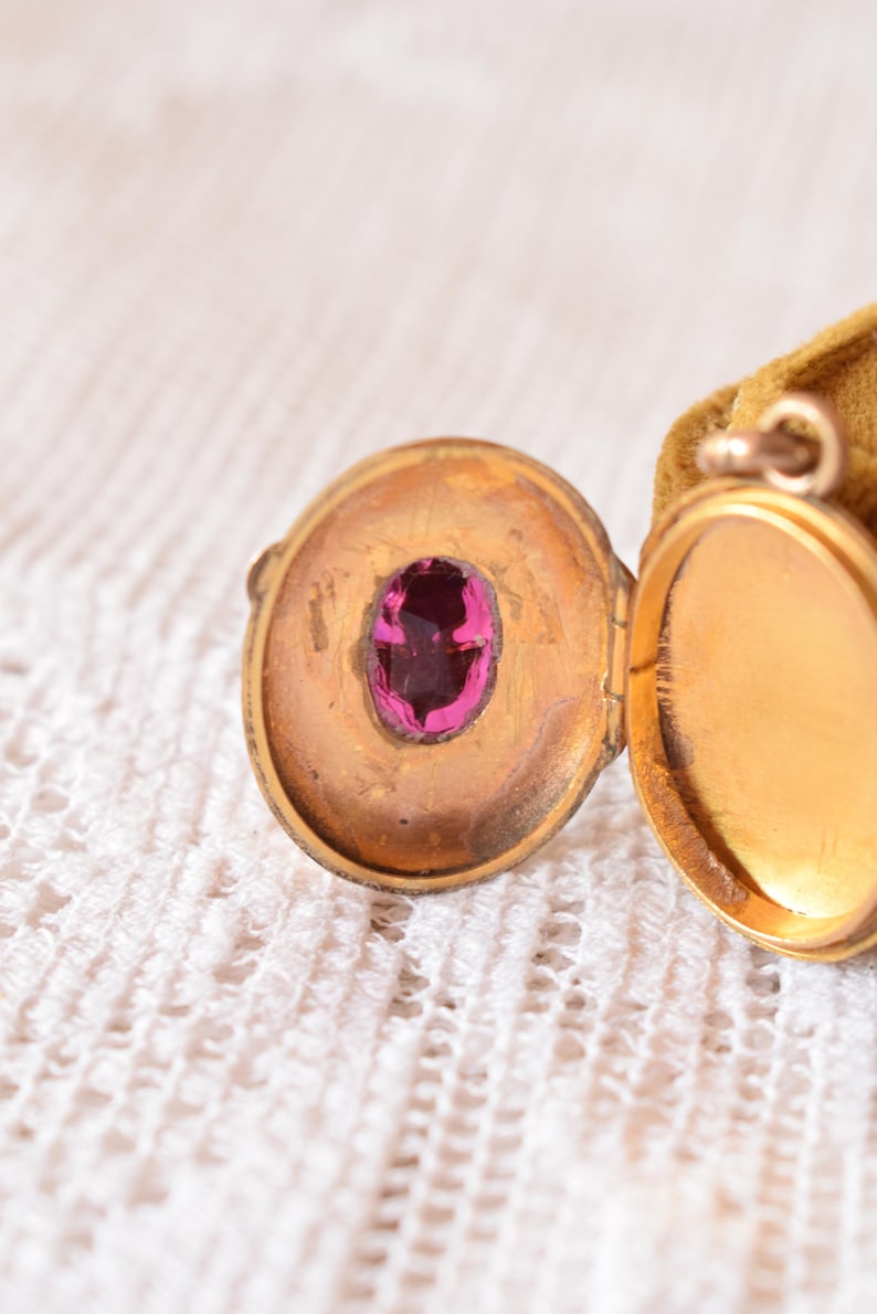Puede incluir: Un relicario ovalado de metal en tono dorado mostrado en posici&oacute;n abierta. El interior del lado izquierdo presenta una peque&ntilde;a piedra preciosa ovalada de color rosa intenso engastada en el centro. El lado derecho del relicario est&aacute; vac&iacute;o y liso. Una peque&ntilde;a anilla met&aacute;lica est&aacute; unida a la parte superior.