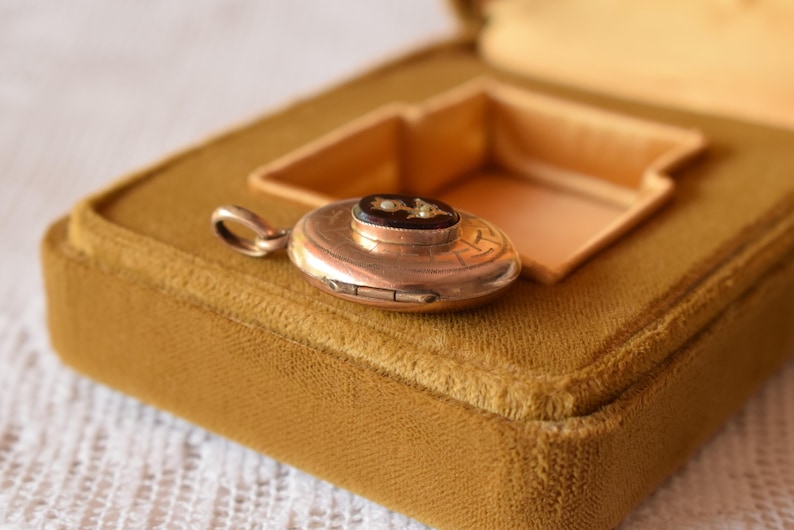 Puede incluir: Un colgante de medall&oacute;n redondo de metal en tono dorado con un patr&oacute;n geom&eacute;trico grabado en la superficie y una incrustaci&oacute;n ovalada negra central decorada con peque&ntilde;os detalles florales blancos. El colgante incluye una peque&ntilde;a anilla circular.