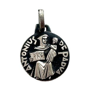 May include: A silver-toned pendant featuring a black and white image of Saint Anthony of Padua holding a book and a baby. The pendant is round and has the words "ANTONIUS DE PADUA" inscribed around the edge.