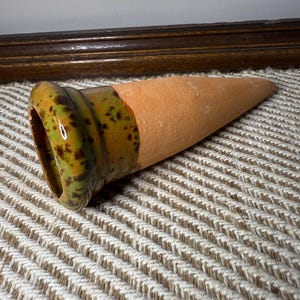May include: A terracotta-colored ceramic plant watering spike with a speckled green and brown rim. The spike is cone-shaped, designed to slowly release water into the soil for potted plants. The object is resting on a textured, light-colored surface.