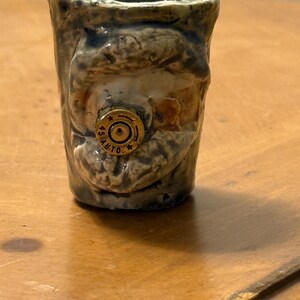 May include: A small, handmade ceramic shot glass with a rustic, textured appearance. The glass is primarily shades of brown and tan, with a gold-colored bullet casing embedded in the side. The casing is marked "45 AUTO".