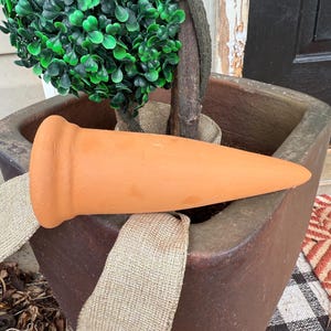 May include: A conical terracotta plant watering spike with a wide, rounded top rim. The object is smooth, matte, and orange-brown in color, featuring a tapered point designed for insertion into soil to provide hydration for potted plants.
