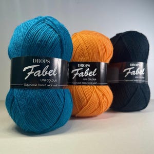 May include: Three skeins of Drops Fabel Uni Colour superwash treated sock wool in teal, orange, and black. Each skein is wrapped with a black label featuring the brand name and product details.