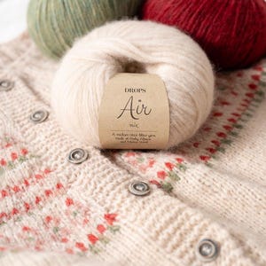 May include: A close-up of a ball of Drops Air yarn with a label, and a cream-colored knitted cardigan with red and green floral patterns. The yarn is a light beige color. The label reads "DROPS Air mix".