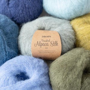 May include: A collection of colorful yarn balls in shades of blue, green, and yellow. The central ball features a brown label with the text "DROPS Brushed Alpaca Silk". The yarn appears soft and fluffy, ideal for knitting or crocheting projects.