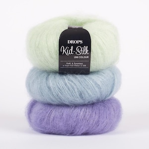 May include: A stack of three balls of Drops Kid-Silk Uni Colour yarn in pastel shades. The top ball is a light green, the middle is a soft blue, and the bottom is a lavender color. A black label is attached to the top ball.