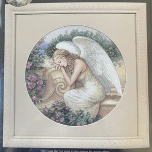 May include: A framed cross-stitch kit from The Gold Collection, featuring a serene angel in a garden setting. The artwork, titled "Serenity's Garden," has a 12-inch diameter. The kit includes a design by Edward Tadiello.