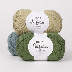 May include: Three skeins of Drops Safran Uni Colour 100% cotton yarn in shades of green, blue, and tan. Each skein has a white label with the brand name and product details. The yarn is tightly wound, suggesting it's suitable for knitting or crochet projects.