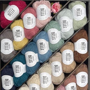 May include: A wooden organizer filled with various colors of Drops Belle yarn. The yarn is in shades of pink, blue, green, yellow, and brown. Each skein has a white label with the text "DROPS BELLE UNICOLOUR". A pink rose is in the background.