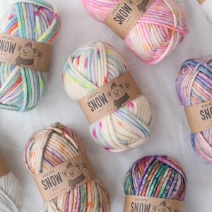 May include: Several skeins of thick, multi-coloured yarn labelled Drops Snow Print. Each ball features a tan paper band with a bear illustration and the text Drops Snow Print 100% Wool. The yarn displays a variegated pattern of pastel pink, blue, yellow, and green hues.