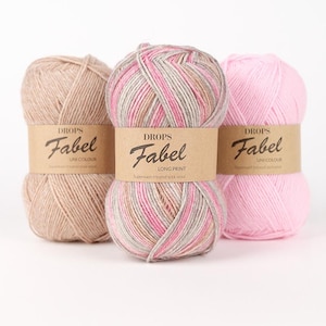 May include: Three skeins of Drops Fabel yarn arranged side by side. The left skein is solid beige, the center skein features a long print of pink, beige, and grey, and the right skein is solid light pink. Each has a brown paper label.