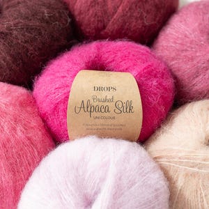 May include: Close-up of several skeins of Drops Brushed Alpaca Silk yarn in various shades of pink and burgundy. A label on one skein reads "Drops Brushed Alpaca Silk UNI COLOUR". The yarn appears soft and fluffy, ideal for knitting or crochet projects.