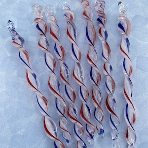 May include: A collection of seven clear glass stir sticks with red and blue spiral accents. Each stick has a decorative top and a twisted design, creating a visually appealing and elegant look. The sticks are arranged on a light blue surface.
