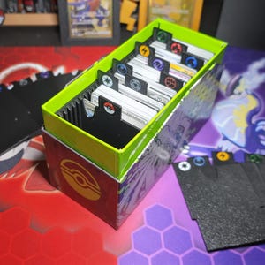 Peut inclure: Une boîte de rangement rectangulaire pour cartes à collectionner avec un intérieur vert vif et un extérieur sombre arborant un logo Poké Ball jaune. La boîte contient plusieurs piles de cartes séparées par des intercalaires noirs avec des icônes colorées.