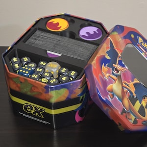 May include: An open, hexagonal Pokémon trading card game tin. The tin features a colorful design with a Charizard illustration. Inside are dice, card dividers, and tokens. The tin has the text "ex" on the side.