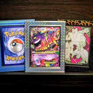 Op de afbeelding: Drie ingelijste Pokémon-ruilkaarten. De kaarten worden weergegeven in blauwe, zilveren en zwarte frames. De kaarten bevatten kleurrijke illustraties van Pokémon-personages en het Pokémon-logo. De achtergrond is donker hout.