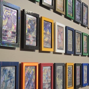 May include: A collection of framed trading cards displayed on a wall. The frames are in various colors, including black, blue, yellow, and orange. The cards feature colorful illustrations and holographic details, creating a visually appealing arrangement.