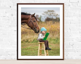 May include: A young girl wearing a blue hat sits on a wooden stool and reads a green book. A brown horse with a bridle looks at the girl from the left side of the image.