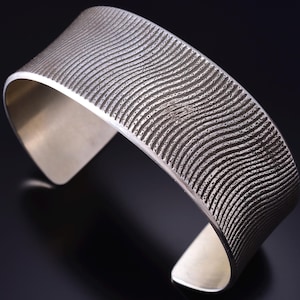May include: A wide, silver cuff bracelet with a textured, wave-like pattern. The open-ended design features a polished interior and a matte, textured exterior. The bracelet is made of a shiny, metallic material.