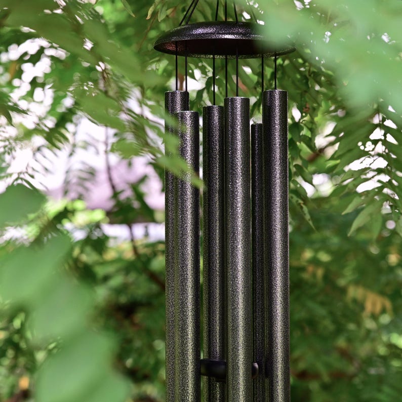 K&ouml;nnte beinhalten: Ein Windspiel mit sechs zylindrischen Metallr&ouml;hren in einer dunklen, texturierten und gesprenkelten Optik. Die R&ouml;hren sind an d&uuml;nnen schwarzen Schn&uuml;ren an einer runden schwarzen Halterung befestigt und bilden eine vertikale Klangstruktur.