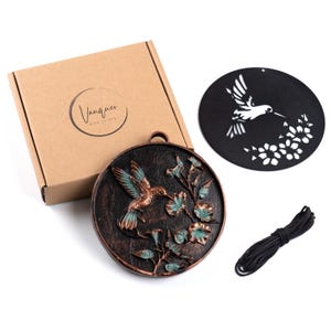 May include: A brown cardboard box with the text Vanquer Wind Chimes, a circular bronze-toned metal plaque featuring a raised hummingbird and floral design with teal accents, a black circular metal cutout of a hummingbird, and a black cord.